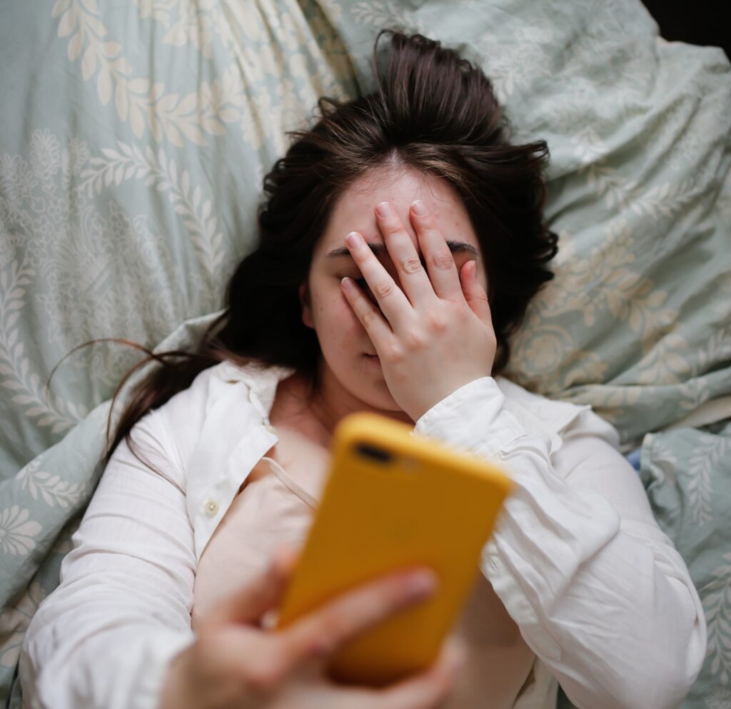 Sleepy young woman ] using phone on bed, close face palm.